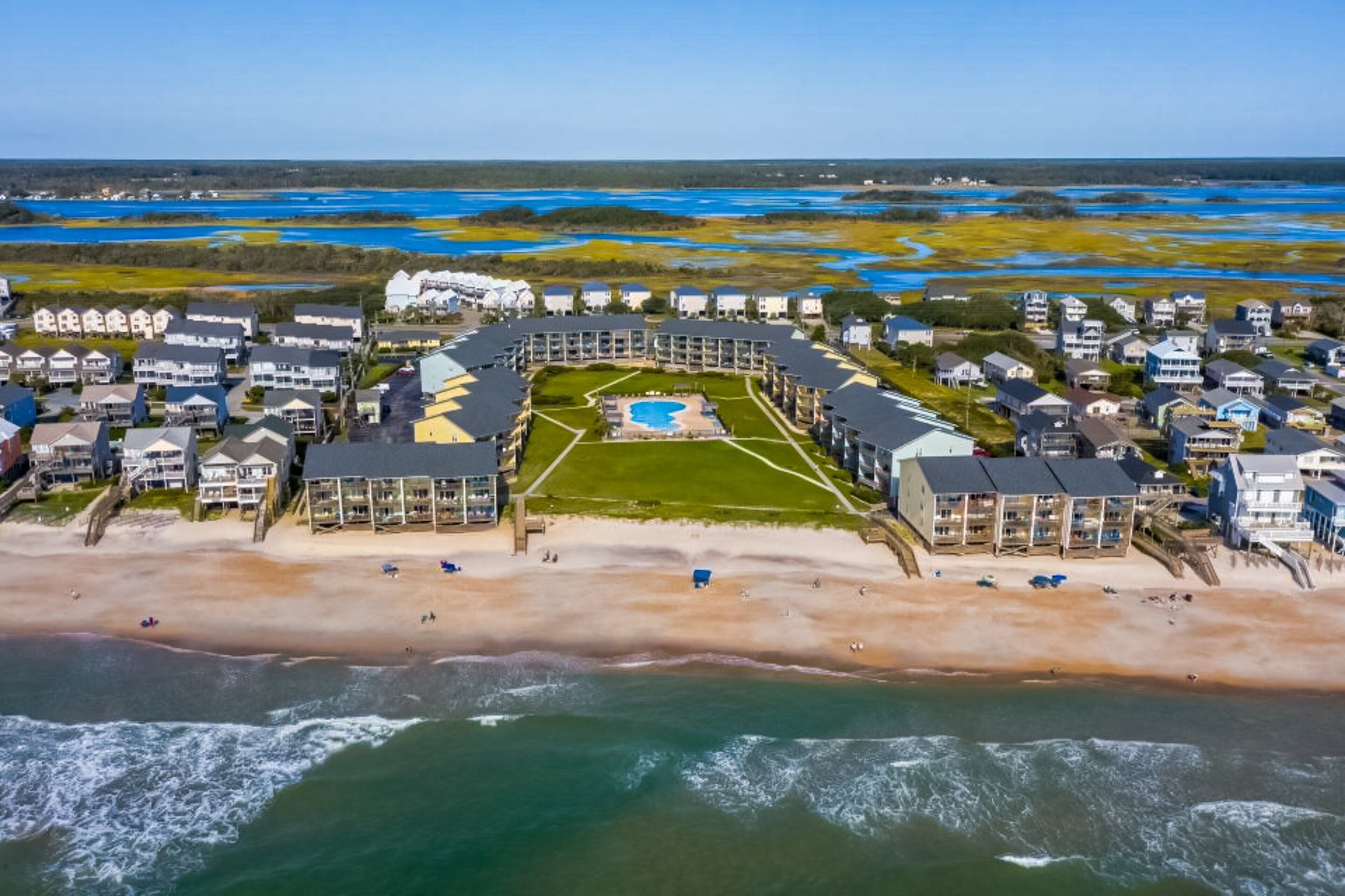An aerial view of a coastal resort with a central pool, beachfront villas, and a sandy beach.