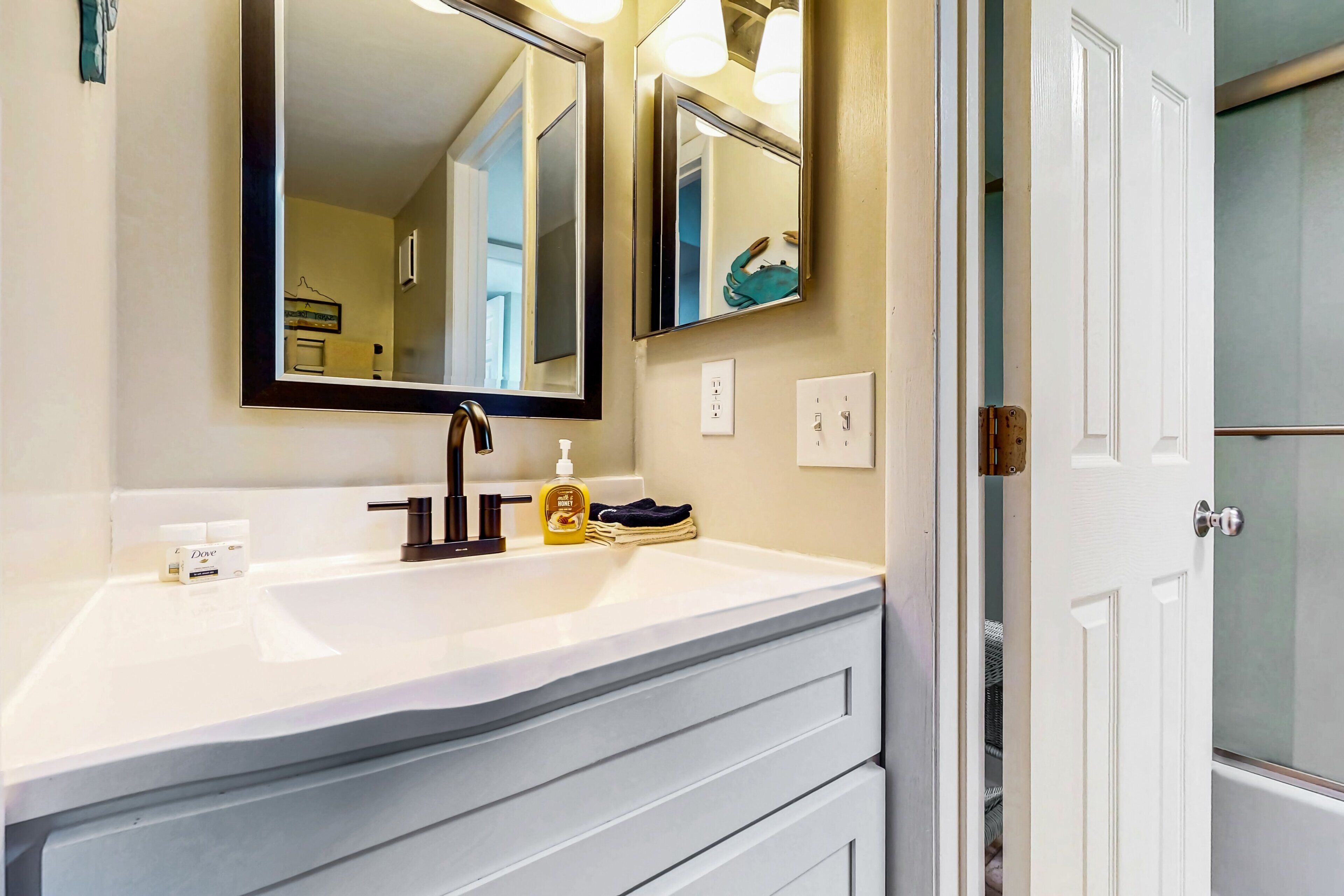 A bathroom with a white sink, two mirrors, a towel rack, and a door leading to another room.