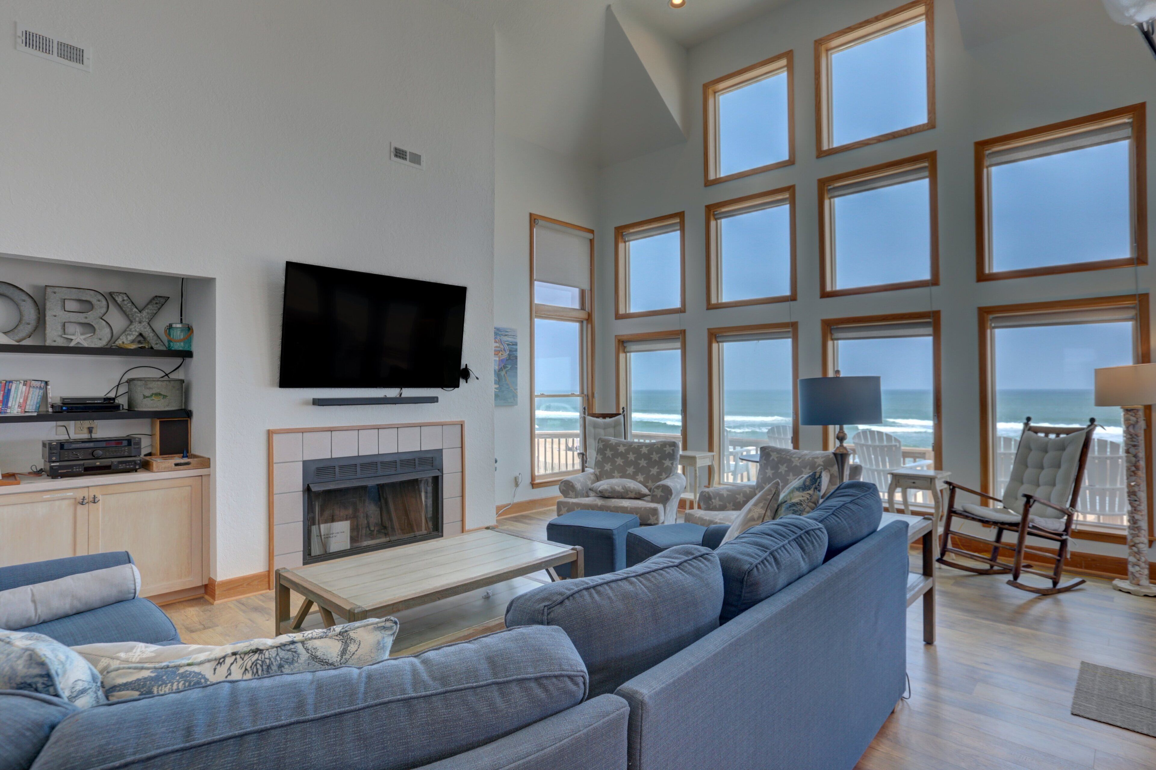 A spacious living room with a large TV, comfortable seating, a fireplace, and a view of the ocean through large windows.