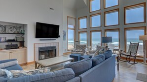 A spacious living room with a large TV, comfortable seating, a fireplace, and a view of the ocean through large windows. - Oceanfront home with hot tub, WiFi, ocean view, central AC, & washer/dryer (Avon)