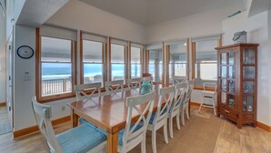 A dining area with a wooden table and white chairs, large windows with ocean views, and a wooden cabinet. - Oceanfront home with hot tub, WiFi, ocean view, central AC, & washer/dryer (Avon)