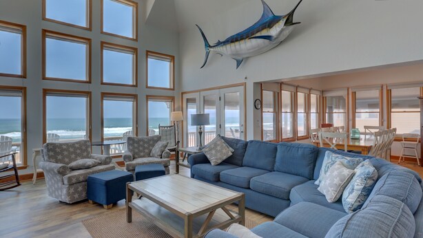 A spacious living room with a large blue sofa, a coffee table, and a view of the ocean through large windows. - Oceanfront home with hot tub, WiFi, ocean view, central AC, & washer/dryer (Avon)