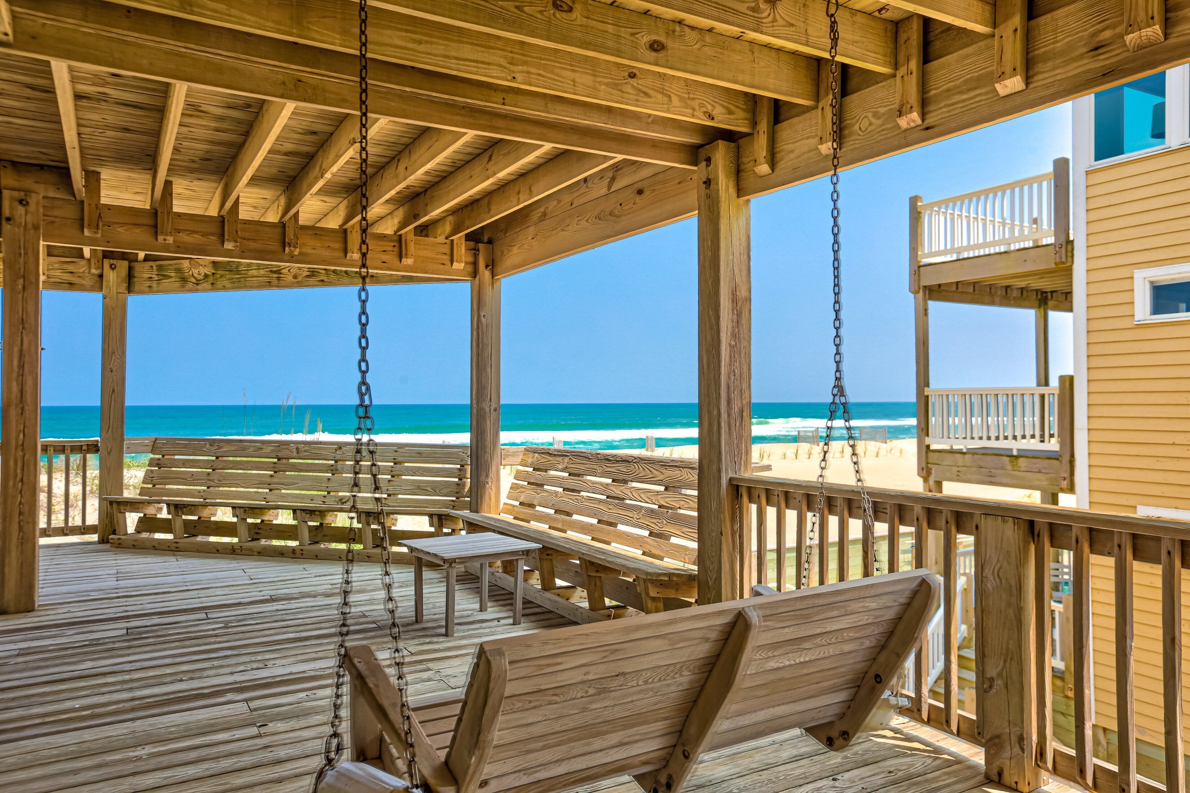 A wooden deck with benches and a swing, overlooking a beach with a clear view of the ocean.
