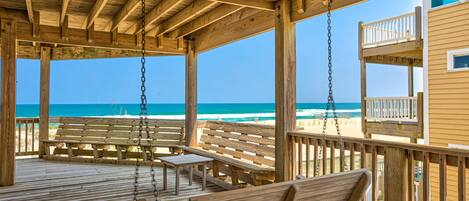 A wooden deck with benches and a swing, overlooking a beach with a clear view of the ocean.