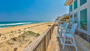 A wooden deck with white Adirondack chairs overlooking a beach with a wooden fence and dunes. - Oceanfront home with hot tub, WiFi, ocean view, central AC, & washer/dryer (Avon)