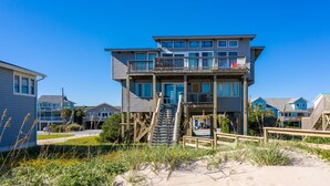Una casa frente al mar de varios pisos, con un balcón y una escalera que conduce al piso superior, y una playa arenosa en primer plano.