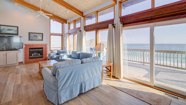 Amplia sala de estar con un sofá grande, una chimenea y un televisor. Grandes ventanas ofrecen vistas a la playa y al océano.