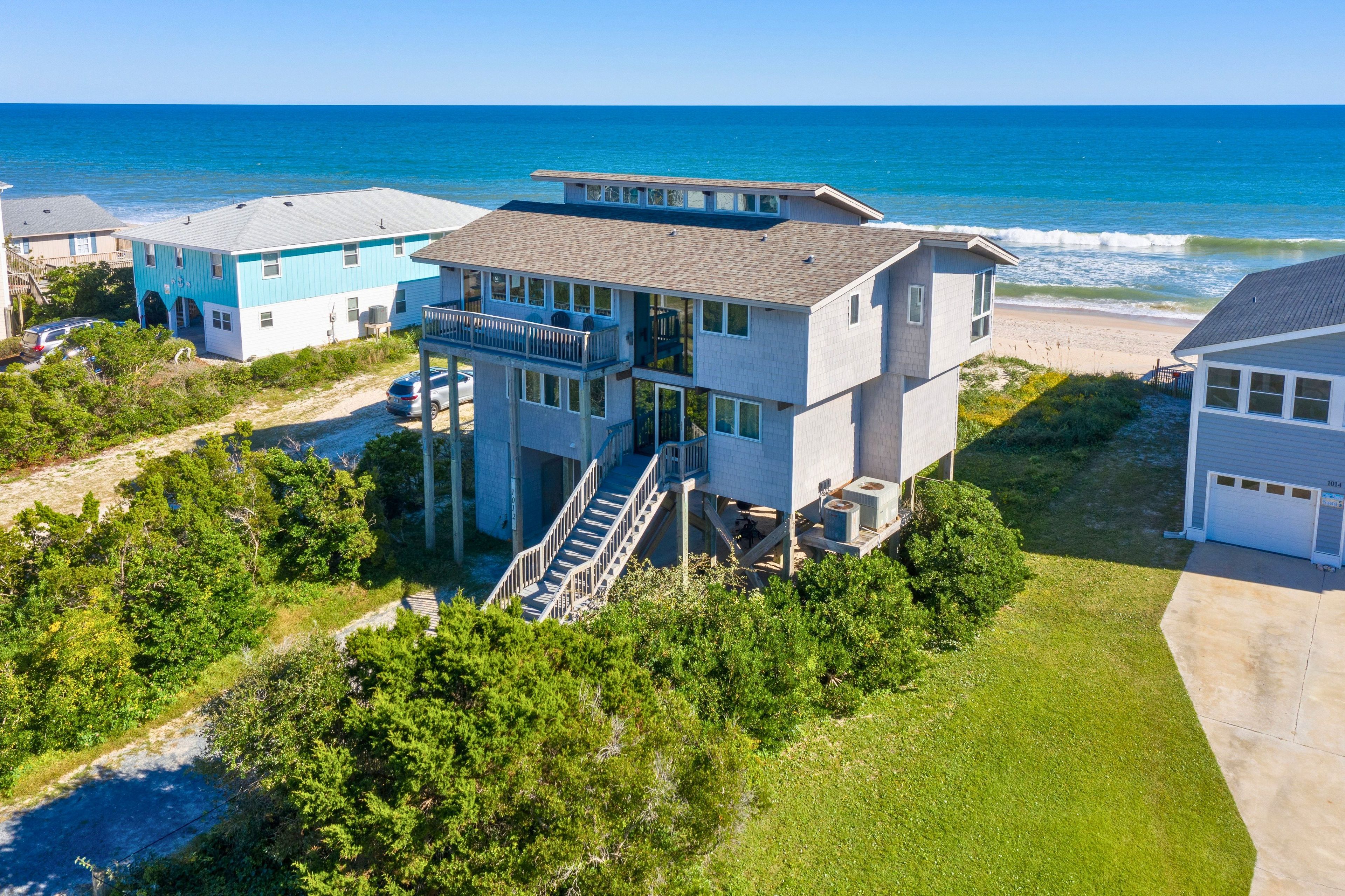 A multi-story beachfront property with a prominent staircase leading to a deck, surrounded by lush greenery and a clear view of the ocean.