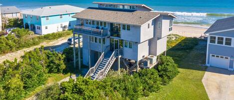 A multi-story beachfront property with a prominent staircase leading to a deck, surrounded by lush greenery and a clear view of the ocean.