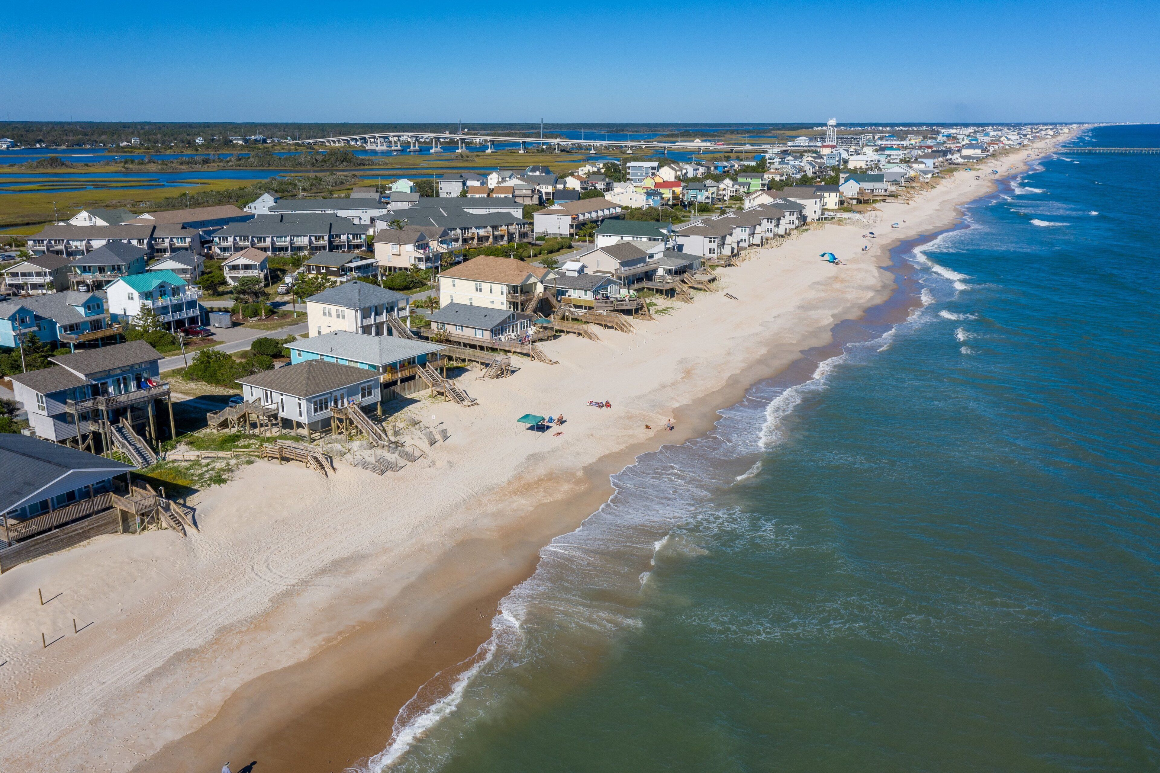 A coastal residential area with houses along the beach, a pier, and a lighthouse in the distance.