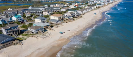 A coastal residential area with houses along the beach, a pier, and a lighthouse in the distance.