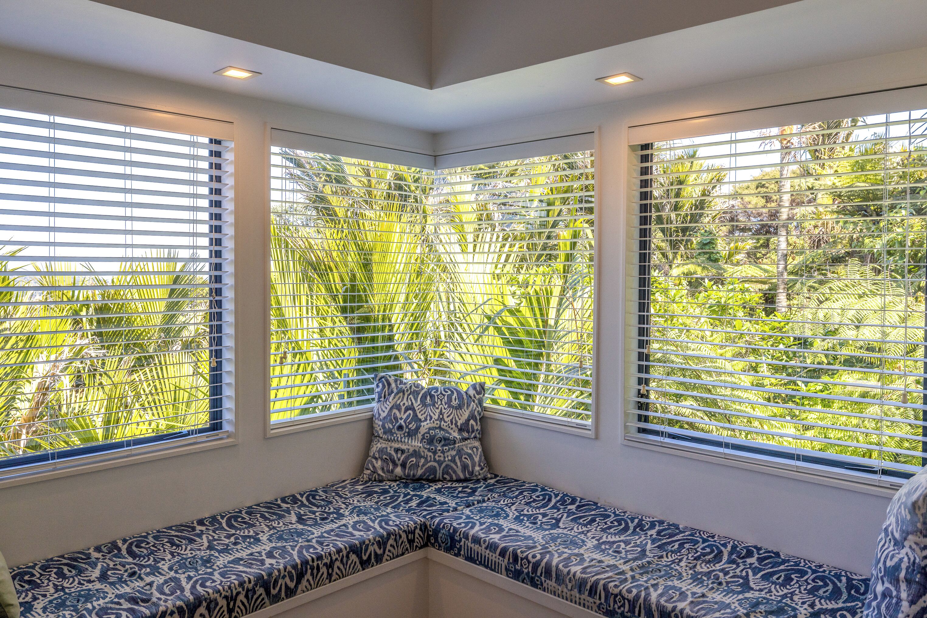 A corner seating area with patterned cushions, large windows with blinds, and recessed ceiling lights.