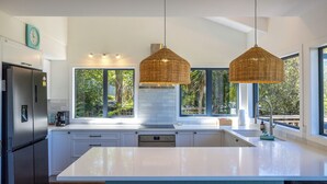 Une cuisine moderne dotée d’un grand îlot central, d’appareils électroménagers en acier inoxydable et de grandes fenêtres offrant une vue sur la verdure.