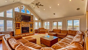 A spacious living room with a large leather sofa, a fireplace, and a balcony with a view of the ocean. - Luxury sound-front home with private pool, hot tub, & dock (Buxton)
