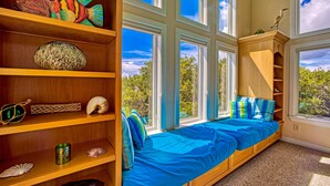 A room with a large window, a bed with blue bedding, wooden shelves with decorative items, and a view of trees outside. - Luxury sound-front home with private pool, hot tub, & dock (Buxton)