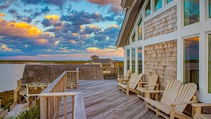 A wooden deck with Adirondack chairs, a view of the ocean, and a modern building with large windows. - Luxury sound-front home with private pool, hot tub, & dock (Buxton)