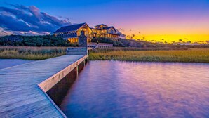 A large, multi-story building with a wooden deck leading to a body of water. - Luxury sound-front home with private pool, hot tub, & dock (Buxton)