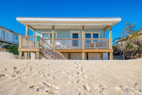 Een huis aan het strand met een houten terras, een trap naar een veranda en een zandstrand op de voorgrond.