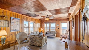 Interior acollidor d'una cabana amb parets de fusta, un ventilador al sostre i vistes a la platja a través de grans finestres.