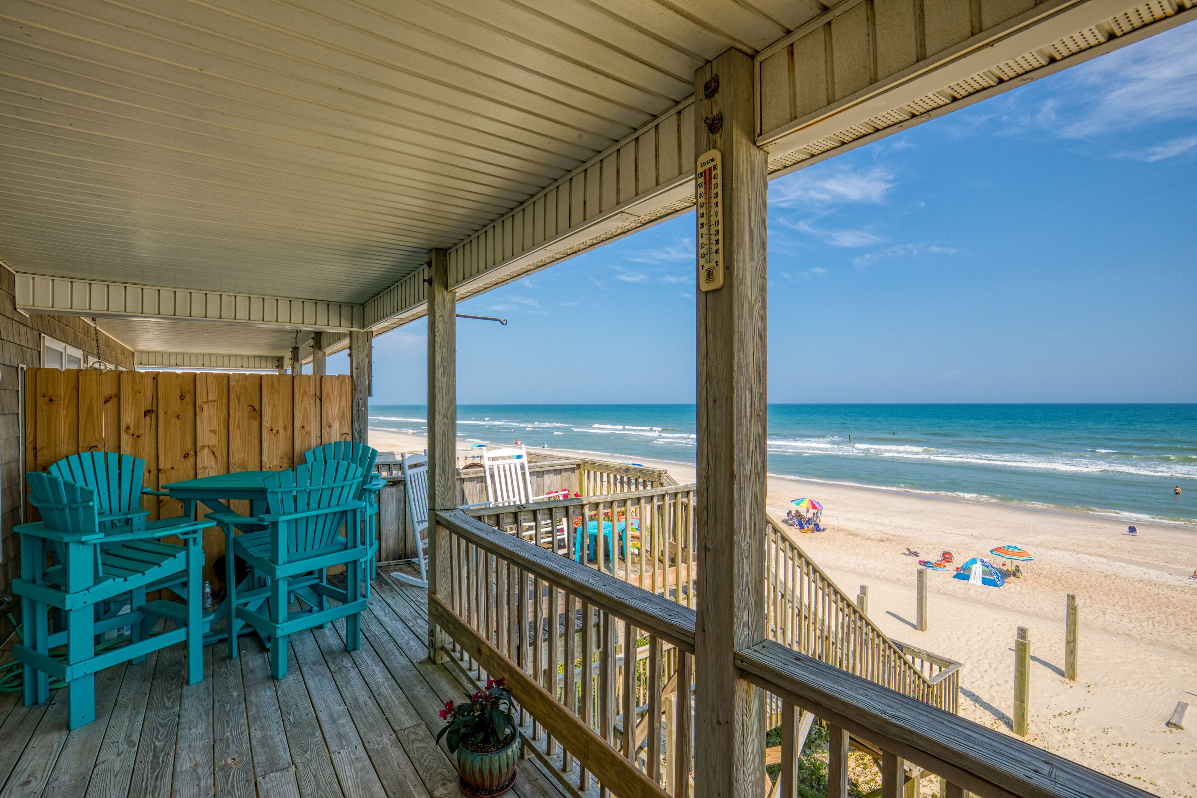 A wooden deck with turquoise Adirondack chairs and a view of the beach.