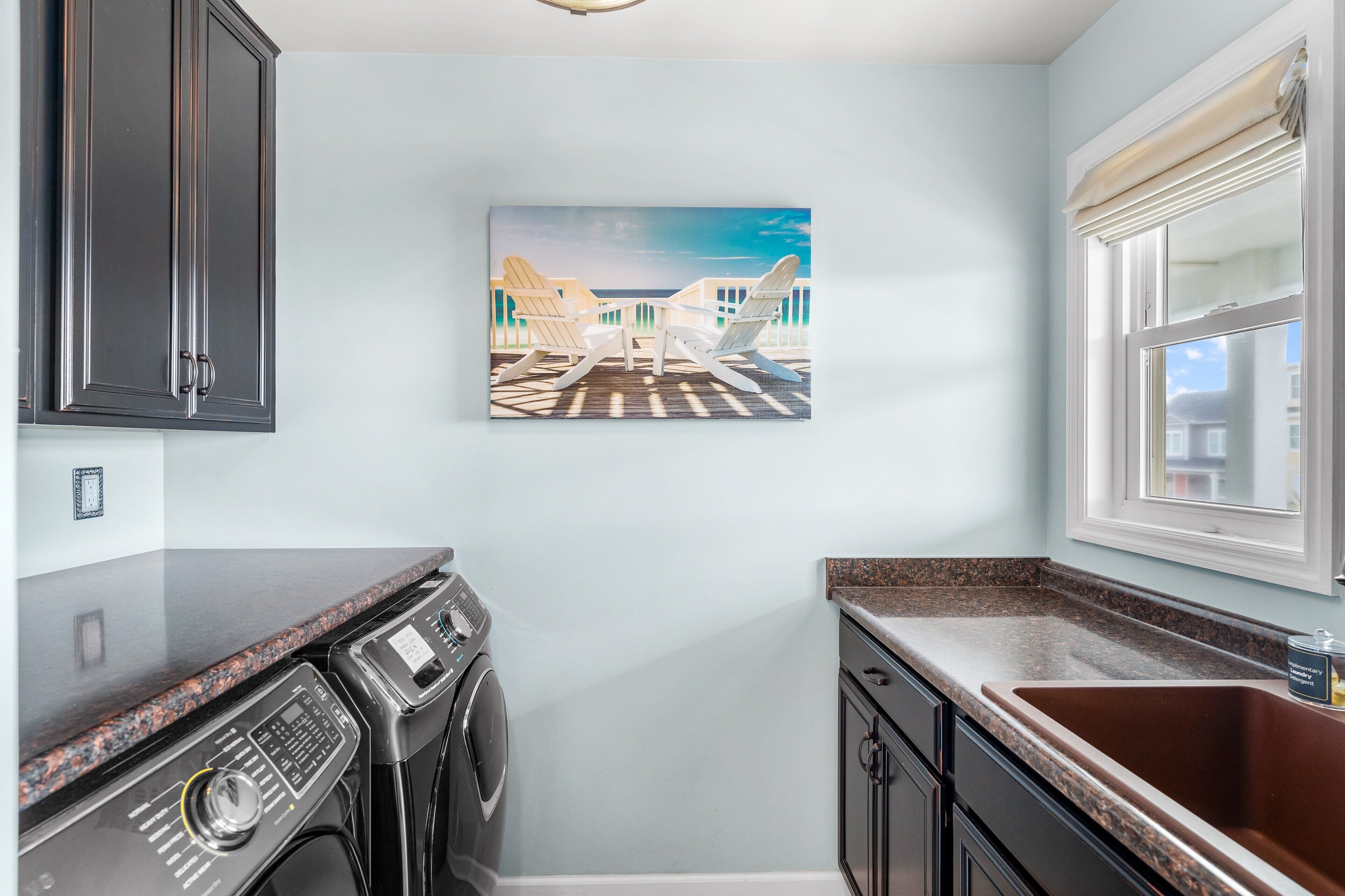 A modern kitchen with dark cabinets, a granite countertop, a built-in washer and dryer, and a large window with a view of the ocean.