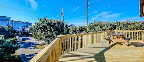Una terraza de madera con una mesa de picnic, rodeada de vegetación y casas al fondo.