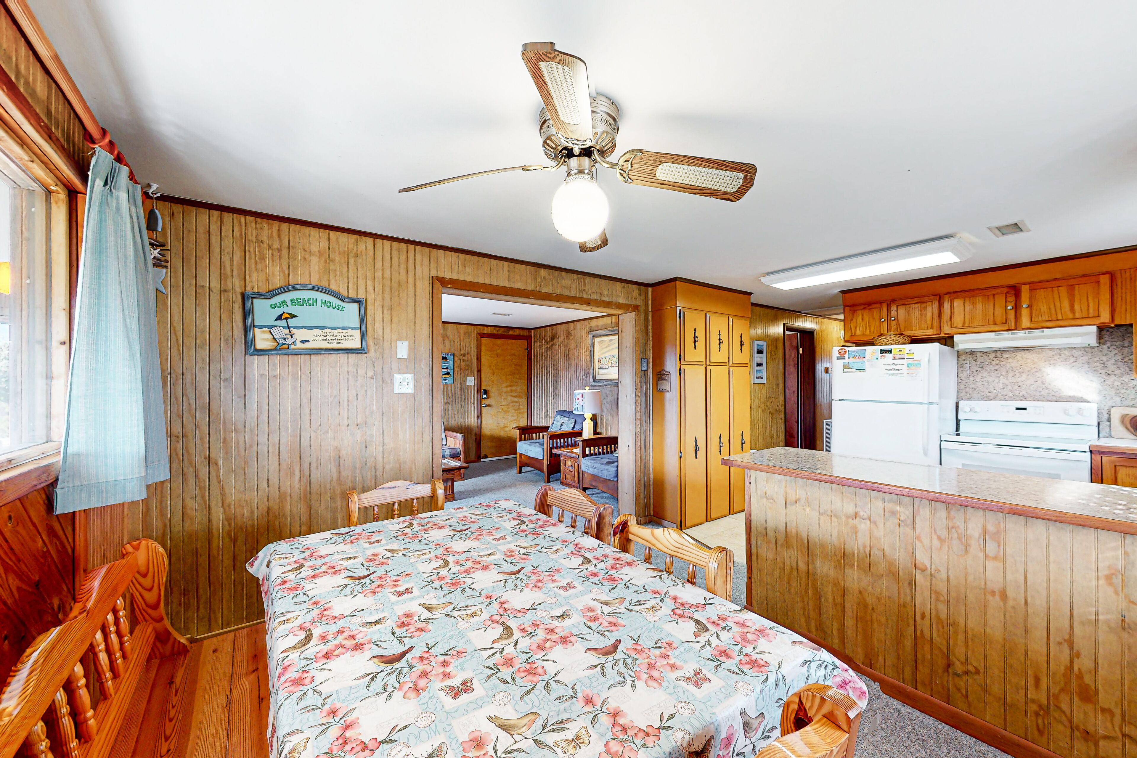 A room with a bed, a ceiling fan, wooden walls, a kitchen area, and a sign that reads 'Our Beach House.'