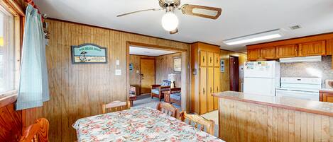 A room with a bed, a ceiling fan, wooden walls, a kitchen area, and a sign that reads 'Our Beach House.'