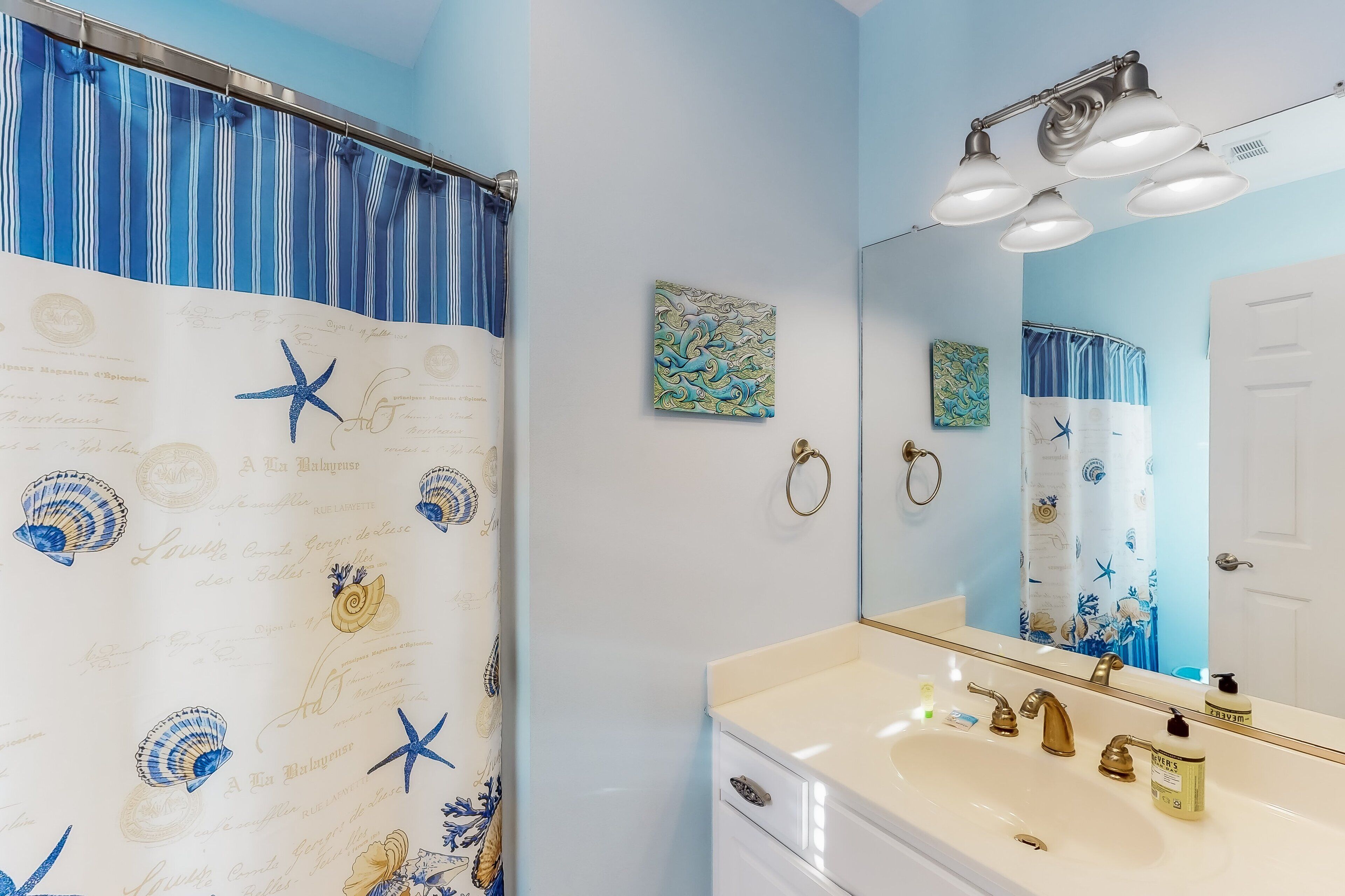 Une salle de bain avec un lavabo blanc, des robinets dorés et un miroir au-dessus. Il y a un rideau de douche à rayures bleues et blanches et orné de motifs d’étoiles de mer.