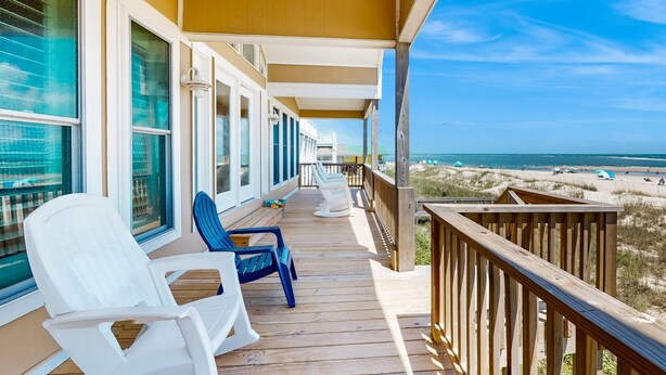 Een houten terras met witte en blauwe stoelen, met uitzicht op een strand met duinen en een heldere lucht.