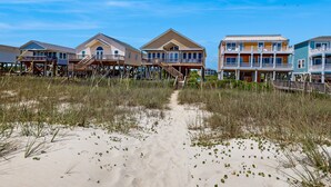 Een pand aan het strand met verhoogde houten constructies, een zandpad dat naar het strand leidt en een helderblauwe lucht.