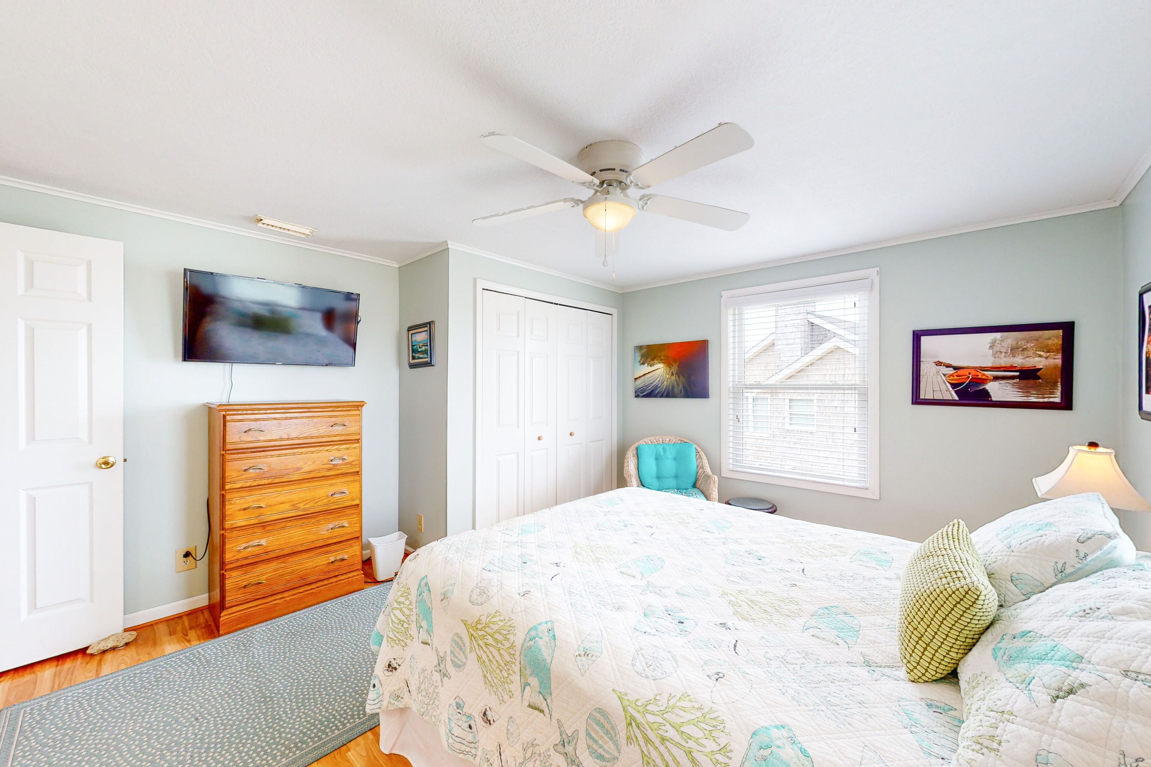 A bedroom with a bed, dresser, ceiling fan, and artwork on the walls.