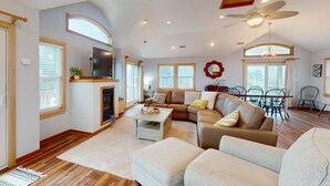 A spacious living room with a fireplace, a large sofa, a coffee table, and a dining area with chairs and a chandelier. - Spacious home with private pool, hot tub, pool table & central AC (Waves)