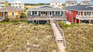 Une maison située sur la côte, dotée d’une terrasse en bois qui mène à un balcon, et entourée de dunes herbeuses.