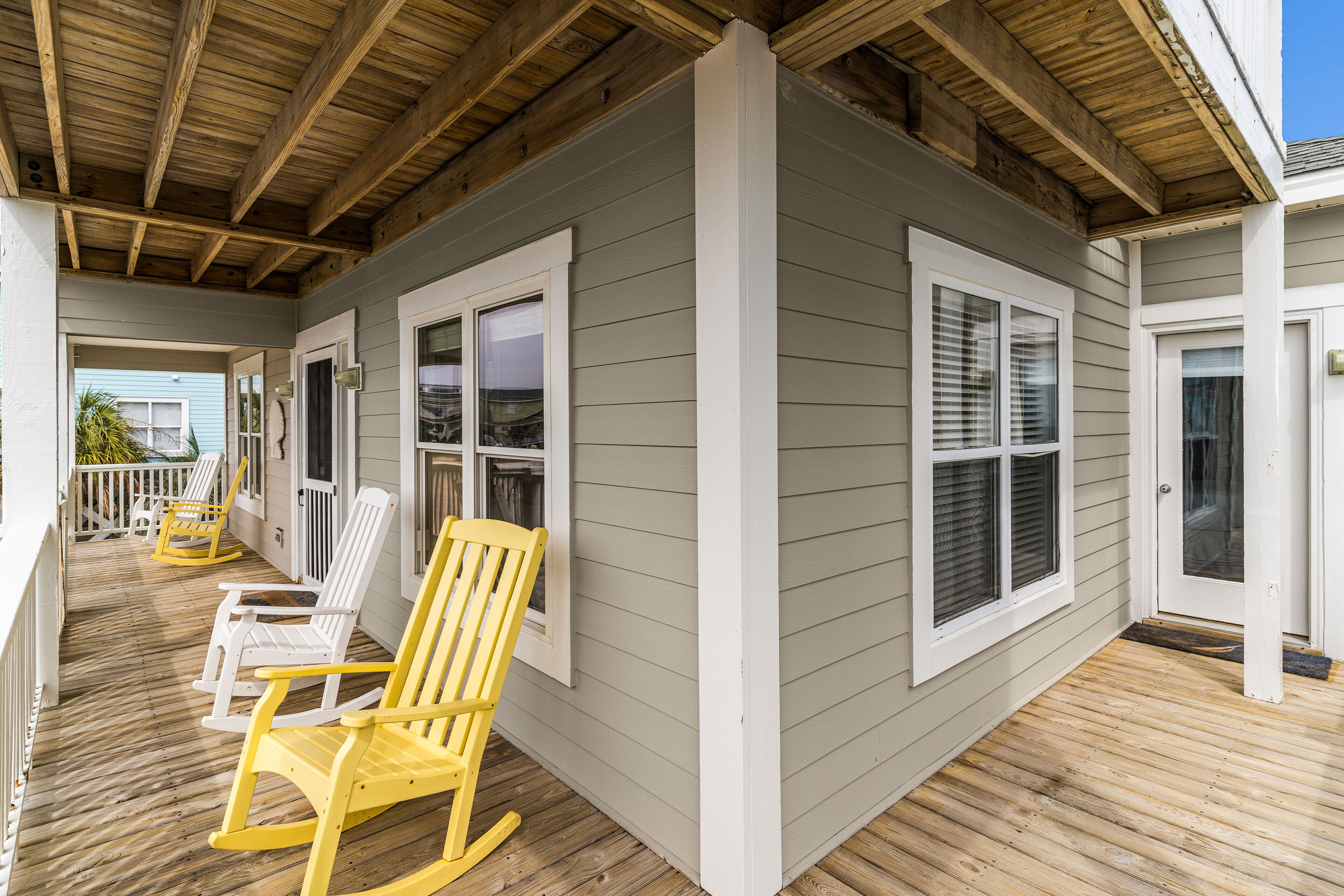 A covered porch with wooden flooring, white and yellow Adirondack chairs, and a grey building with white trim.