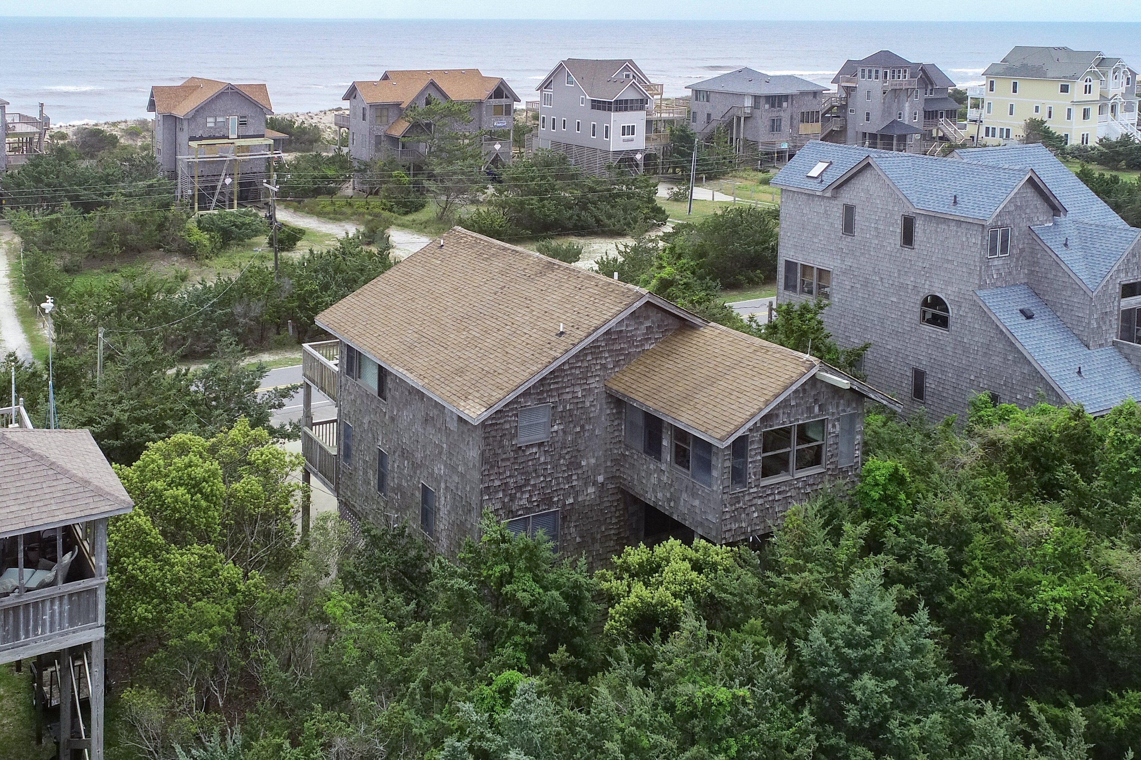 A coastal residential area with houses featuring stone and wood exteriors, surrounded by dense greenery and a view of the ocean.