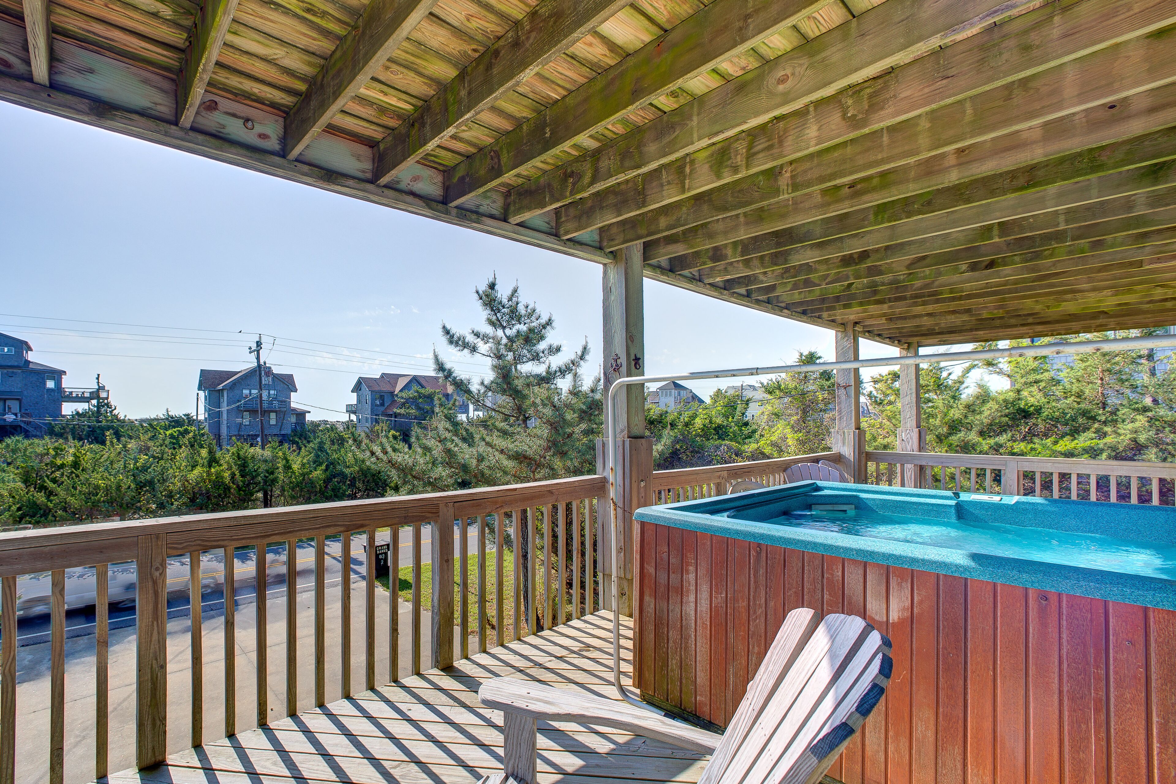 Une terrasse en bois avec un jacuzzi et une chaise, donnant sur une rue bordée de maisons et d’arbres.
