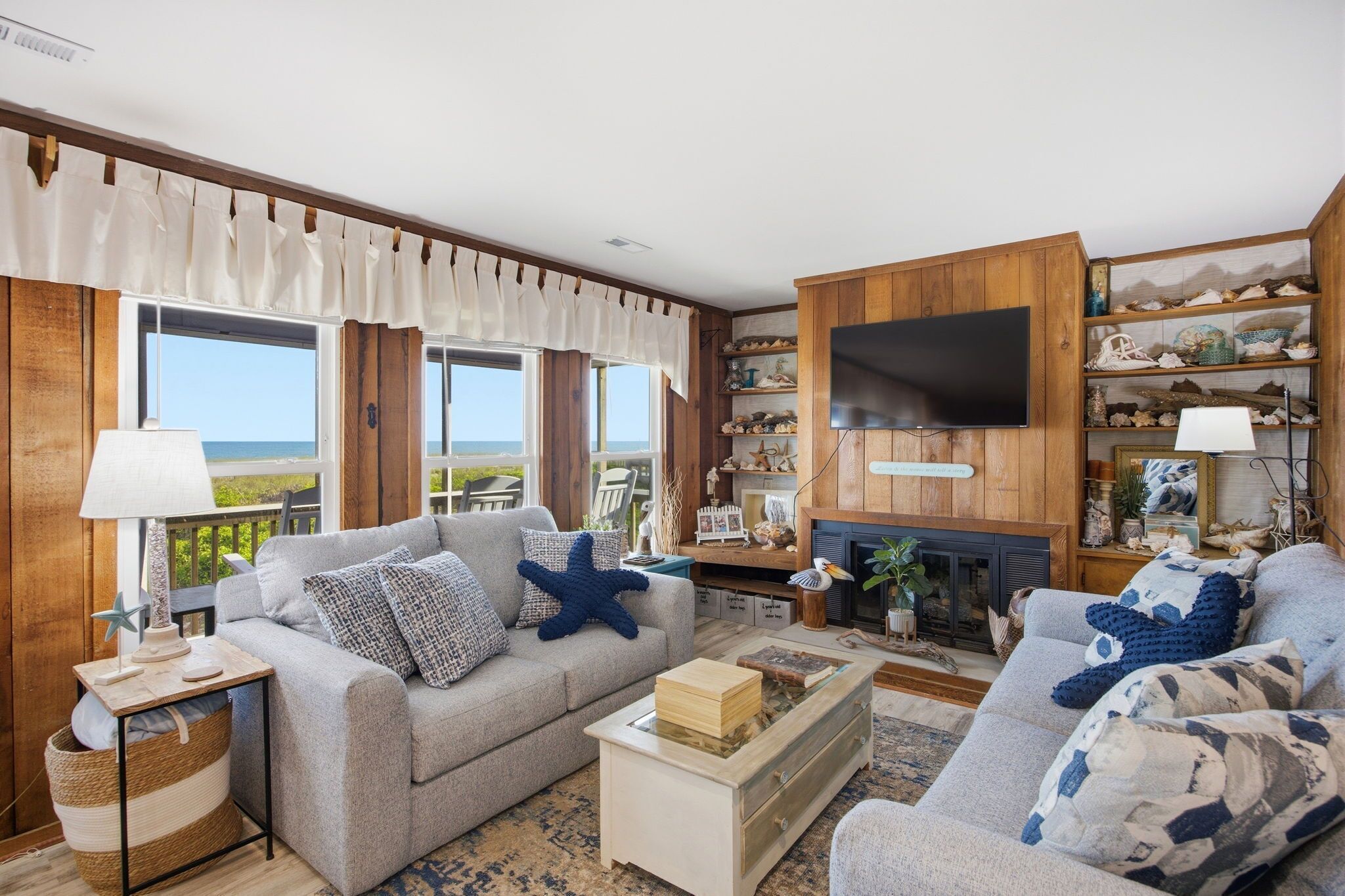 A living room with a sofa, coffee table, and a fireplace. There is a television mounted on the wall and a window with curtains.