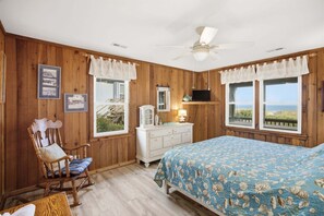 Un dormitorio con paredes de madera, una cama con colcha estampada, un sillón mecedora, un aparador y un ventilador de techo.