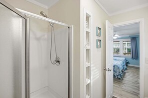 Une salle de bain avec une douche à parois de verre, une porte blanche et un couloir menant à une chambre à coucher équipée d’un lit et d’une fenêtre.