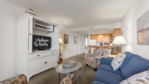 Ein Wohnzimmer mit Fernseher, einer Couch und gemusterten Sesseln, im Hintergrund eine Küche und ein Essbereich mit Tisch und Stühlen.