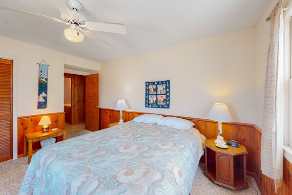 A bedroom with a bed, two bedside tables with lamps, a ceiling fan, a wooden door, and a wall decoration.