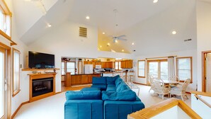 Amplia sala de estar con un sofá seccional azul grande, una chimenea y un televisor. Se ve la cocina al fondo, con armarios de madera y una zona de comedor con sillas blancas.