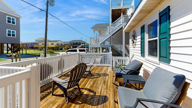 Une terrasse en bois avec des sièges d’extérieur, une clôture blanche et un escalier menant à un balcon.