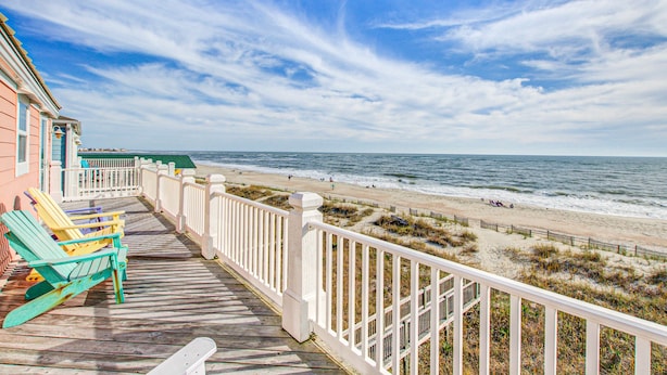 Eine Holzterrasse mit bunten Adirondack-Stühlen mit Blick auf einen Strand, der von einem weißen Zaun gesäumt wird und einen Meerblick bietet.