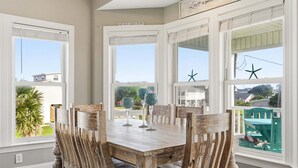 Een eetgedeelte met een houten tafel en stoelen, grote ramen met decoratieve zeesterren en uitzicht op een groen buitengebied.