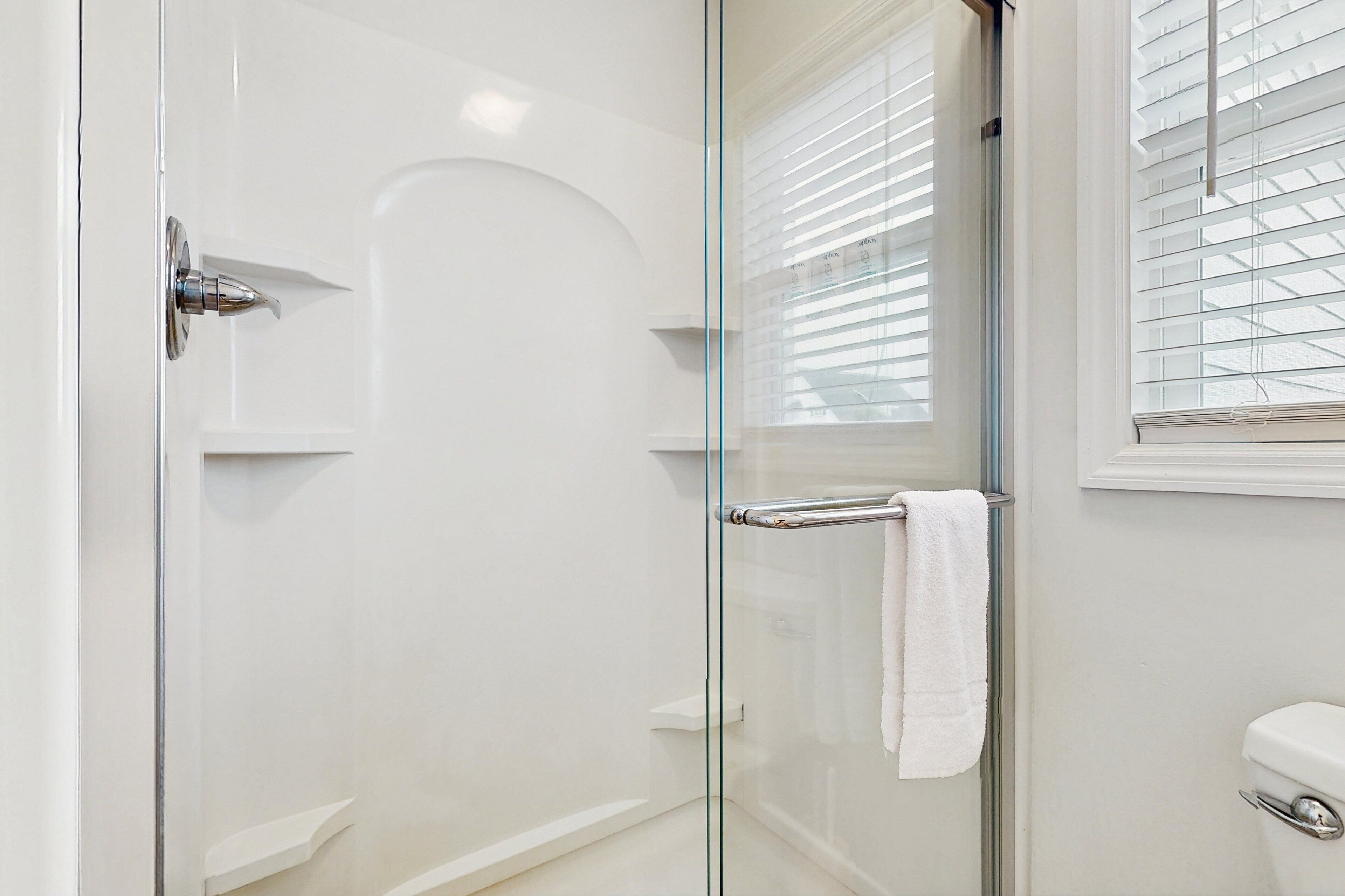 A modern bathroom with a glass shower enclosure, a white toilet, and a window with blinds.