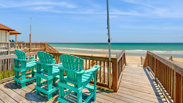 Eine Holzterrasse mit türkisblauen Adirondack-Stühlen mit Blick auf einen Strand.