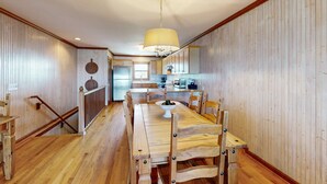 Ein Esszimmer mit Holzboden, einem großen Tisch und Stühlen, einer Hängelampe und einer Küche im Hintergrund.