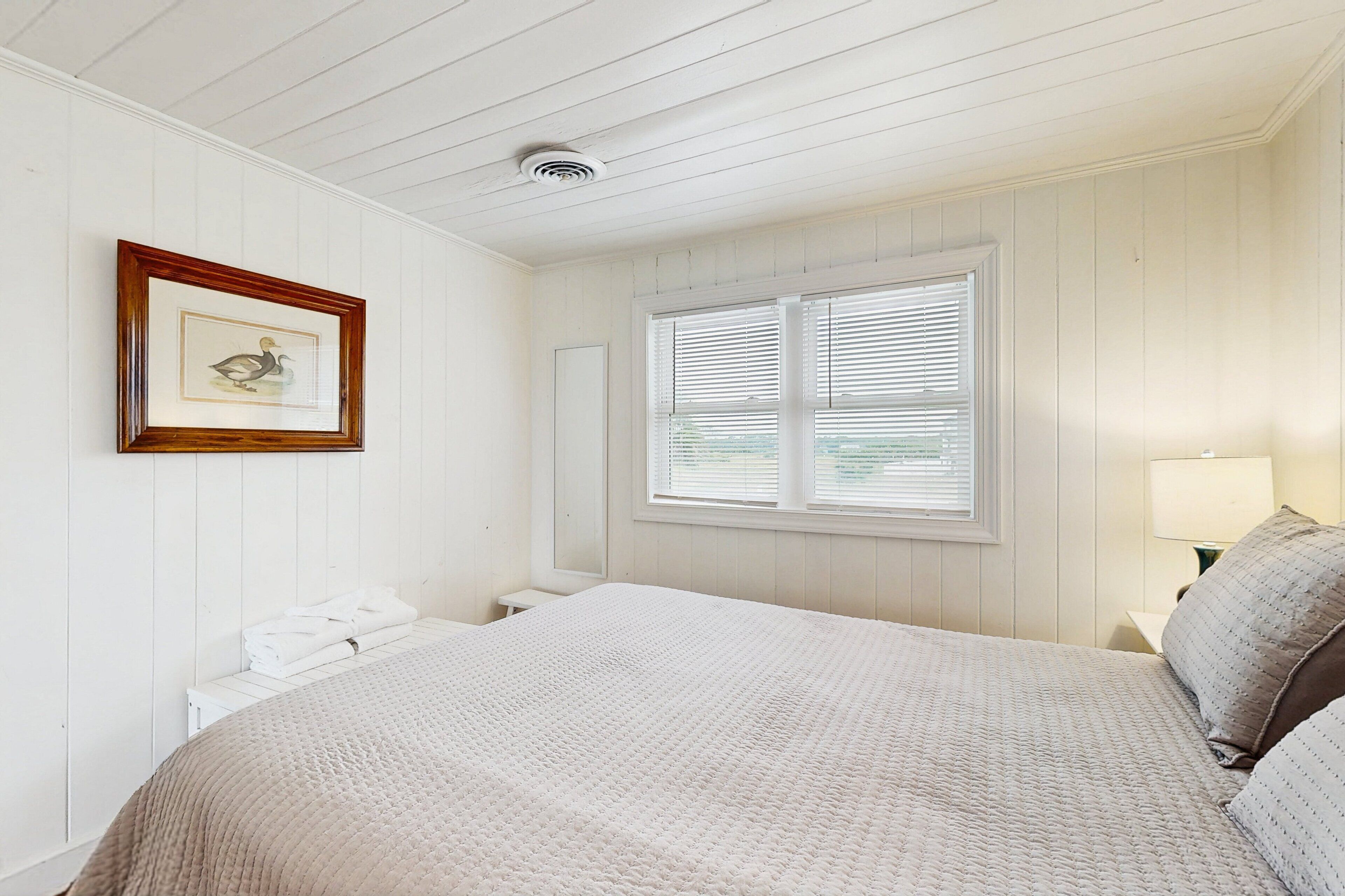 Un dormitorio con cama, un cuadro en la pared, una ventana con persianas y una mesita de noche con lámpara.